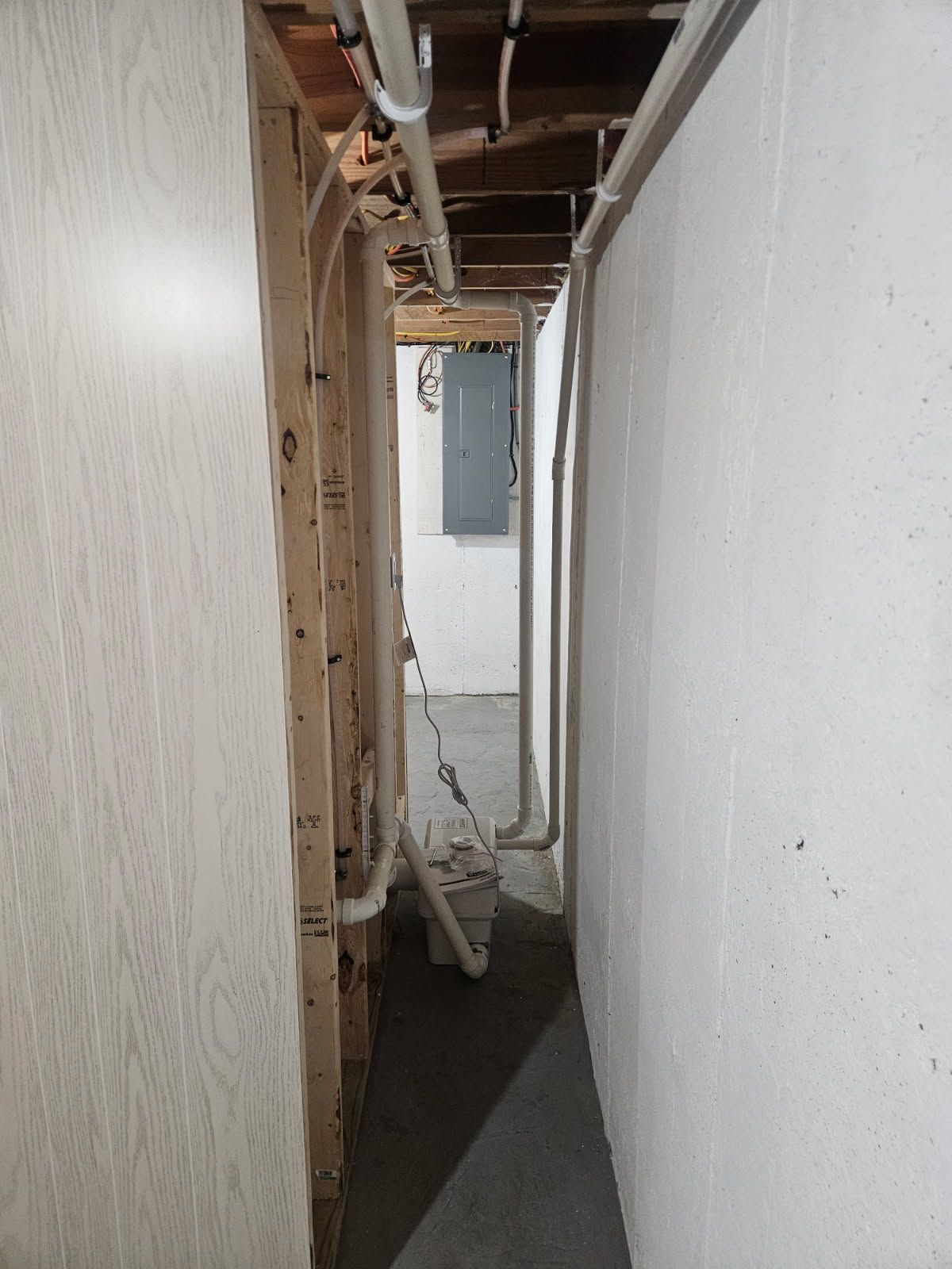 A narrow, unfinished basement hallway with exposed wood framing, white walls, and overhead plumbing pipes.