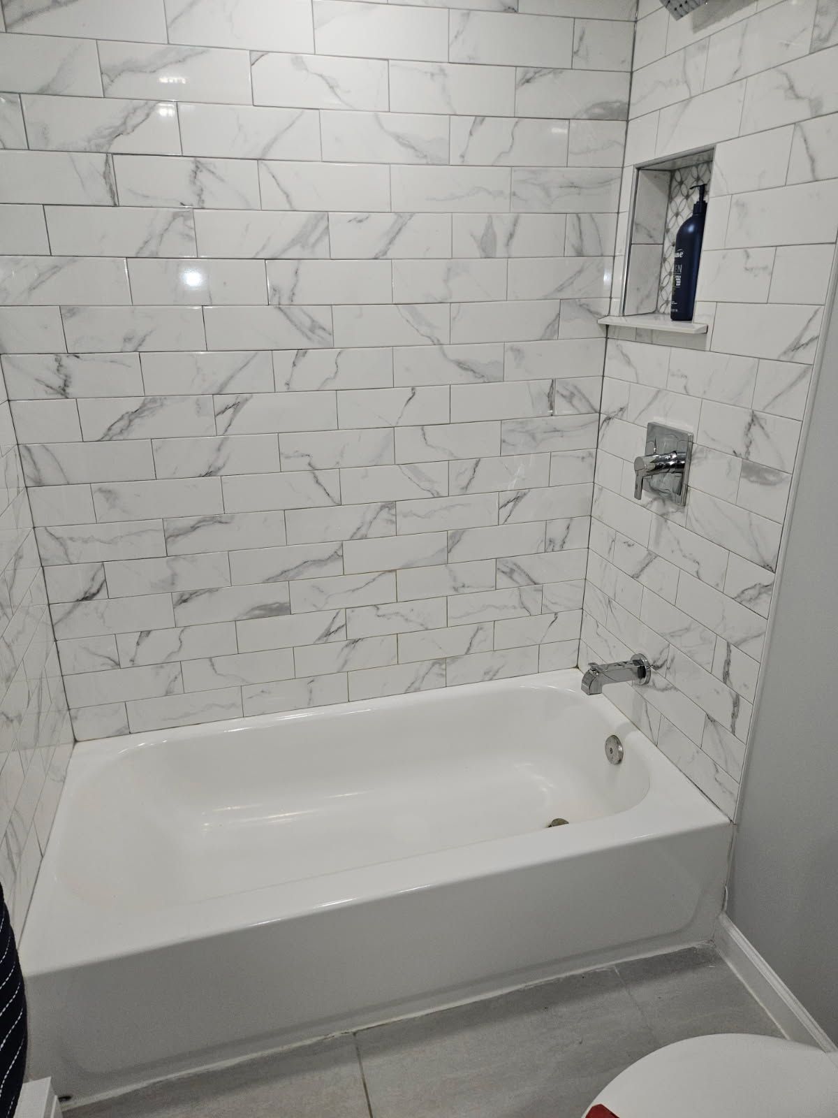 A white bathtub with marble-look subway tile walls, a built-in recessed shelf, and chrome fixtures.