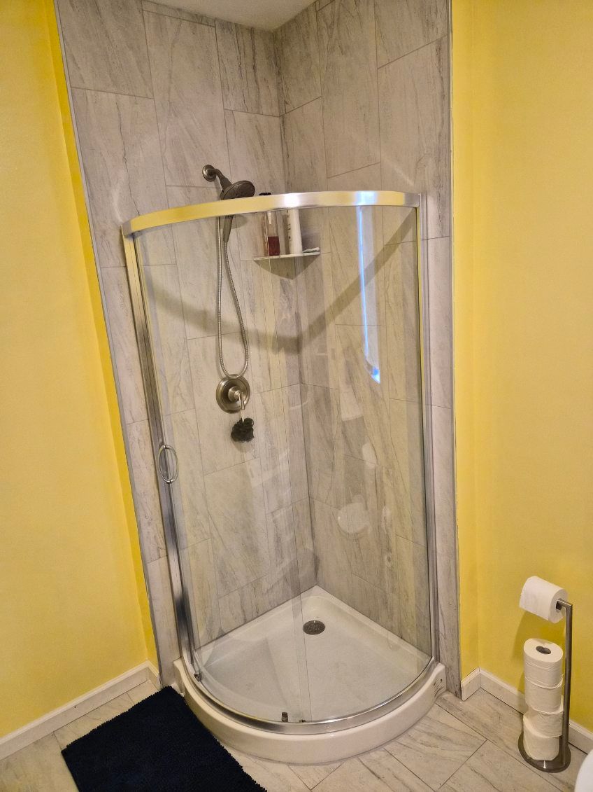 A corner shower stall with a curved glass door and gray stone-look tiles, situated in a bathroom with yellow walls.