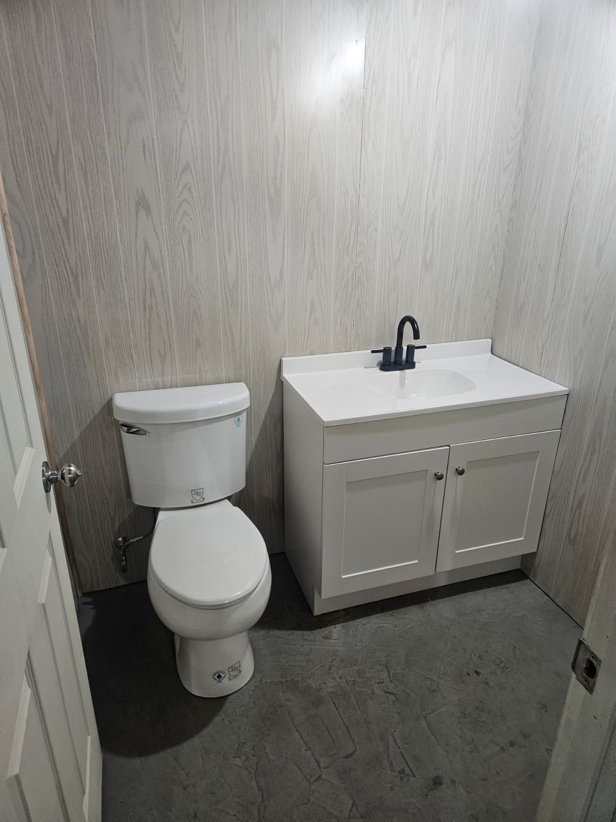 A small bathroom with a white toilet and a white vanity with a black faucet against light, wood-paneled walls.