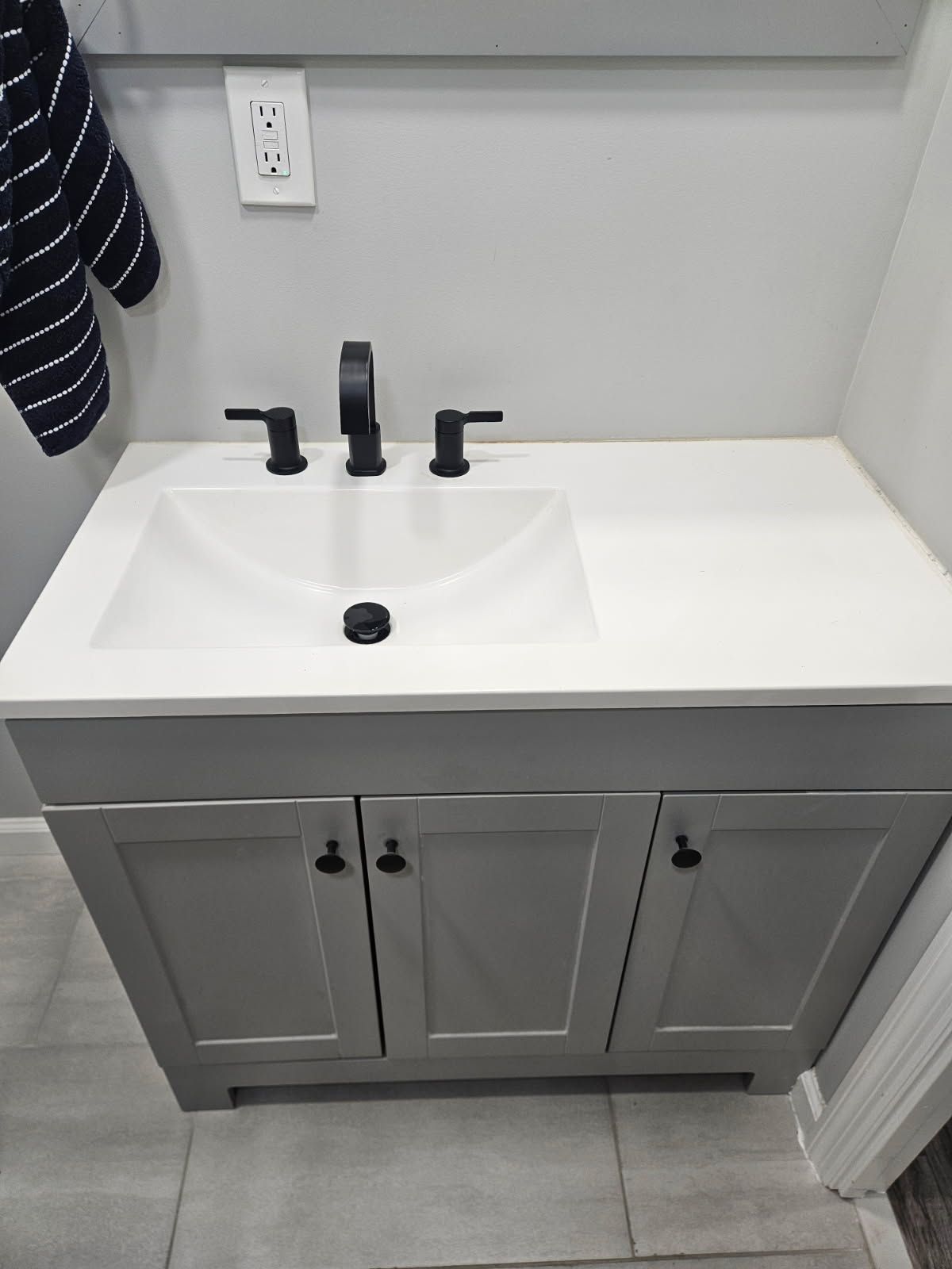 A modern bathroom vanity with a white rectangular sink, matte black faucet, and gray cabinets on a tiled floor.