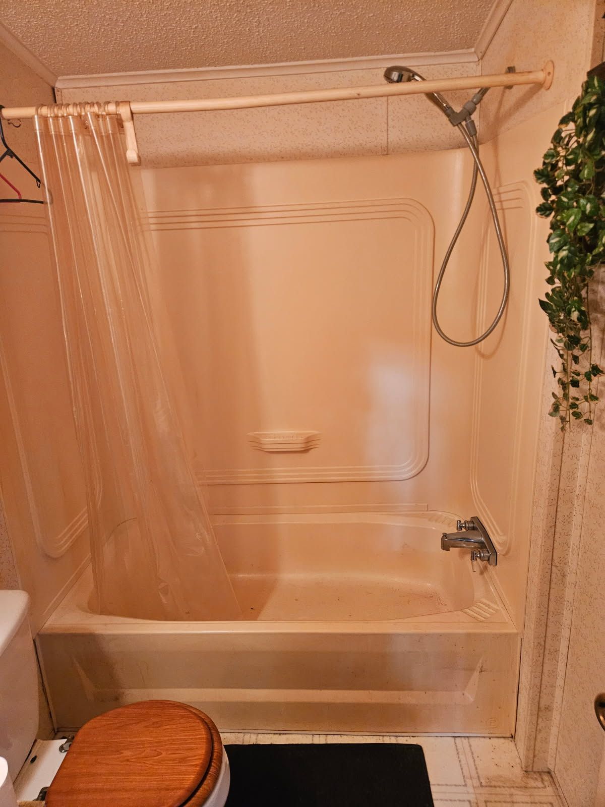 A beige fiberglass bathtub and shower stall with a clear curtain, a hand-held shower head, and a toilet in the foreground.