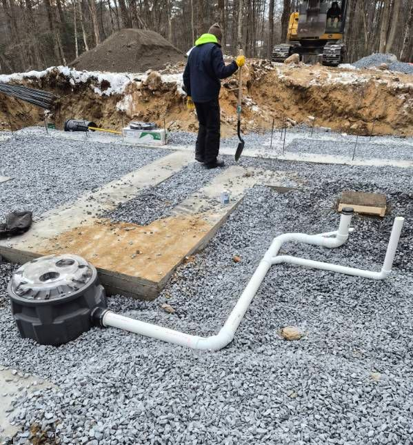 A person holds a shovel on a gravel construction site with white PVC pipes and a black sump pump basin in the foreground.