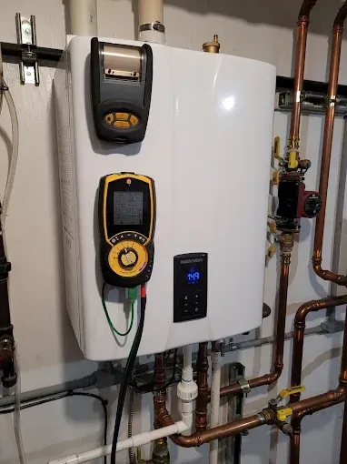 A white tankless water heater mounted on a wall with a digital diagnostic tool and printer attached to its front.