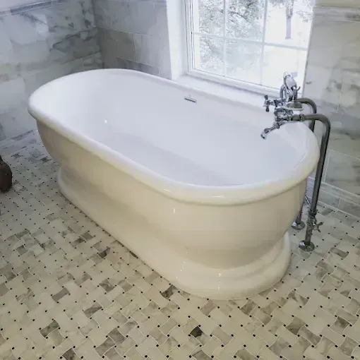 A white, oval freestanding bathtub with a chrome faucet stands on a patterned, light-colored tile floor by a window.