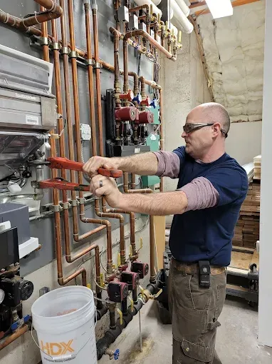 A worker in safety glasses and a navy shirt uses a large wrench to repair copper pipes on a wall in a mechanical room.