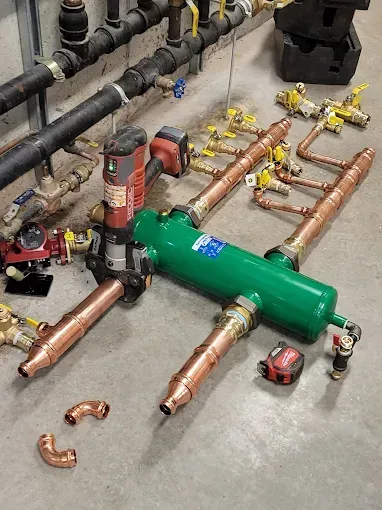 A green hydraulic separator lying on a concrete floor next to copper plumbing pipes, tools, and fittings.