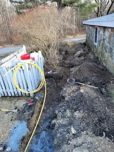 A coiled yellow utility line sits next to a trench dug along a stone building and a white picket fence in a yard.