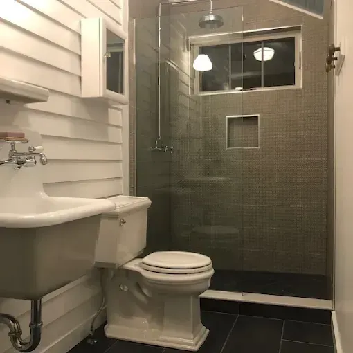 A small bathroom featuring a vintage-style wall-mounted sink, a white toilet, and a glass-enclosed shower with dark tiles.