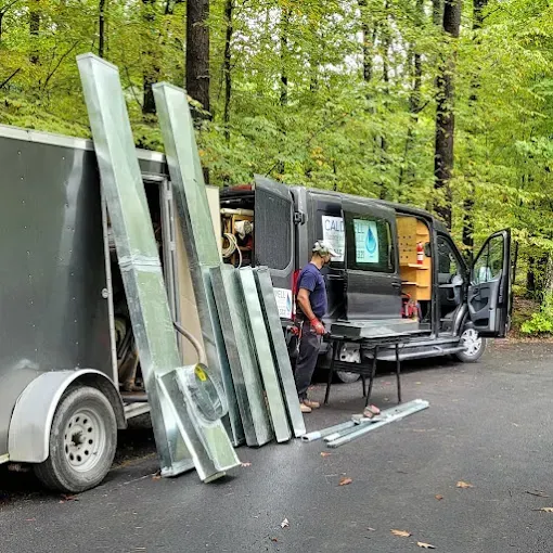A worker stands by a van and trailer filled with metal ductwork in a wooded, outdoor area.