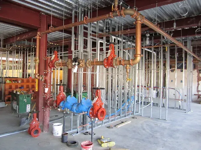 Construction site interior with exposed metal wall framing, copper overhead pipes, and several large red valves.