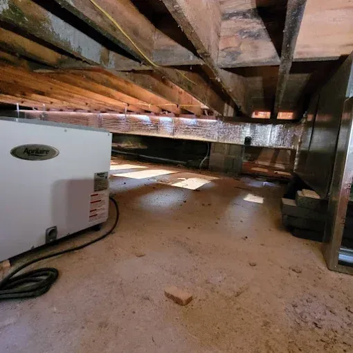 A white ApriAire unit sits in a crawl space with exposed wooden floor joists and silver ductwork overhead.