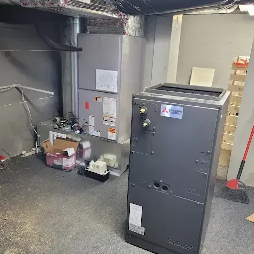 Two HVAC units stand in a basement: a tall, light-gray furnace in the back and a dark-gray Mitsubishi air handler in front.