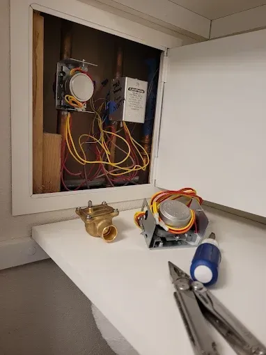 An open wall access panel revealing interior wiring and two zone valves, with tools and a spare valve on a white shelf.