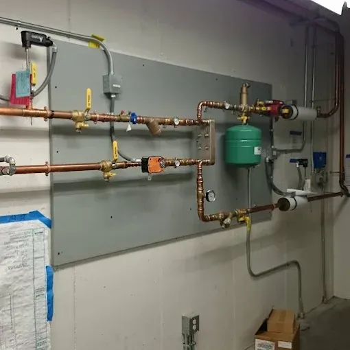 A wall-mounted plumbing system with copper pipes, valves, a green expansion tank, and electrical control components.