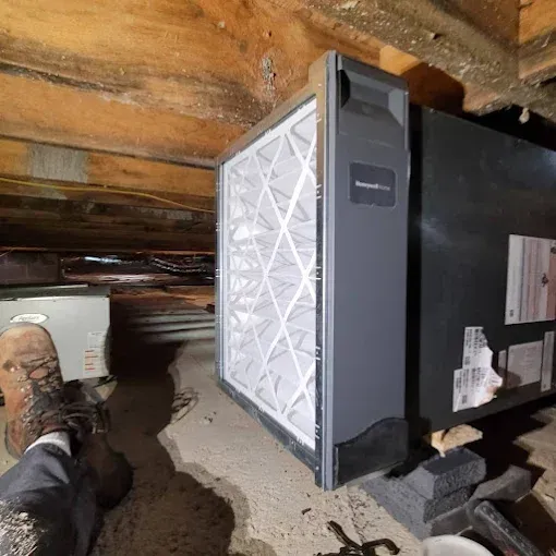 A person’s booted foot rests near a large HVAC air filter cabinet mounted to a furnace in a crawl space.
