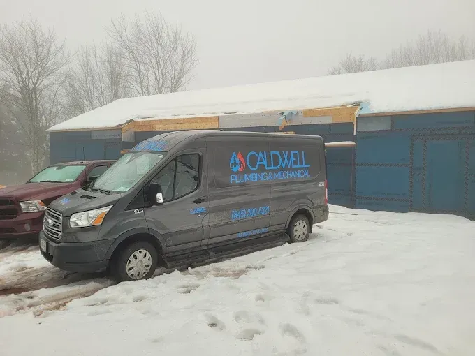 A grey Caldwell Plumbing & Heating service van parked in a snow-covered lot next to a blue building on a cloudy day.