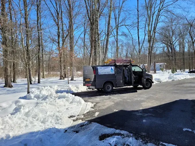 A dark grey van with accessibility signage parked on a paved lot beside a snowy, wooded area on a bright sunny day.