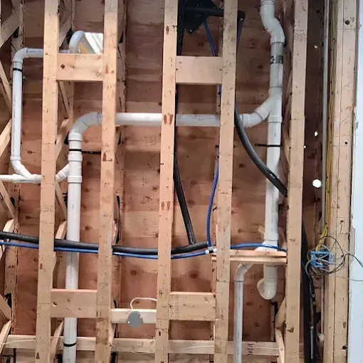 White PVC plumbing pipes run vertically and horizontally through exposed wooden wall studs.