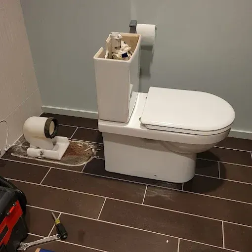 A white toilet with an open tank sits on dark tile next to a detached toilet floor flange in a room with blue walls.