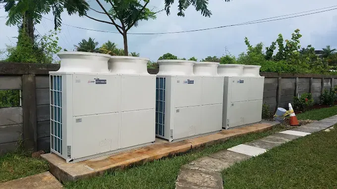 Three commercial outdoor HVAC condenser units sit on a concrete pad against a wall in a grassy yard.