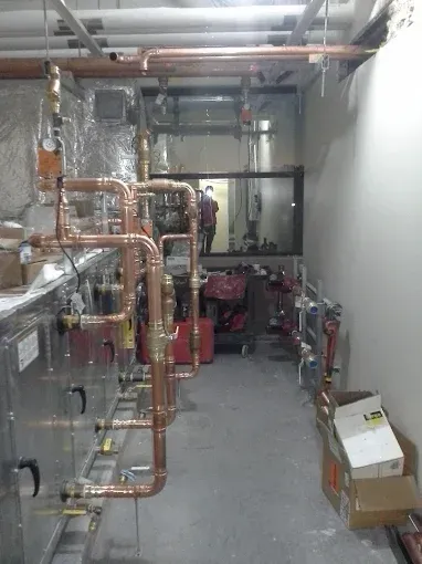 A mechanical room with copper plumbing pipes connected to industrial HVAC equipment, showing valves and metal ductwork.