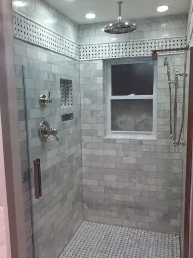 A walk-in shower with light grey marble subway tiles, a rain showerhead, a hand sprayer, and a small window.