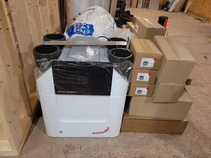A Zehnder heat recovery ventilation unit sits on a floor next to several stacked cardboard boxes in a construction space.