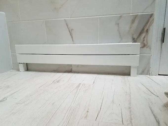 A white, horizontal radiator cover sits on a light-colored tile floor against a wall with marble-patterned tiles.