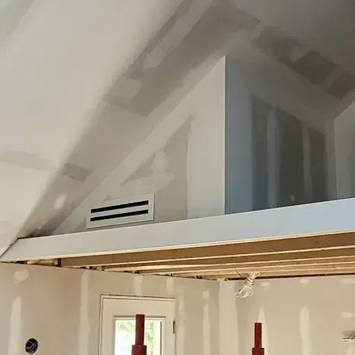 A low-angle view of a room under construction, showing a glossy white finished ceiling above an exposed wooden frame.