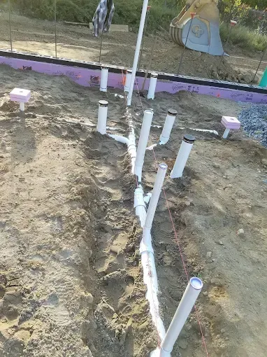 White PVC plumbing pipes installed in a trench at a construction site before a concrete slab is poured.