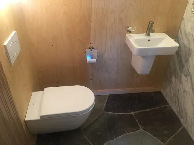 A small bathroom featuring a wall-mounted white toilet and sink against plywood walls, with dark, irregular stone flooring.