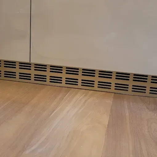 A gold-colored, louvered ventilation panel installed at the base of light-colored kitchen cabinets on a wooden floor.