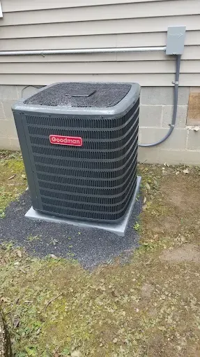A grey Goodman brand outdoor HVAC condenser unit sits on a gravel pad against a vinyl-sided house with a grey utility box.