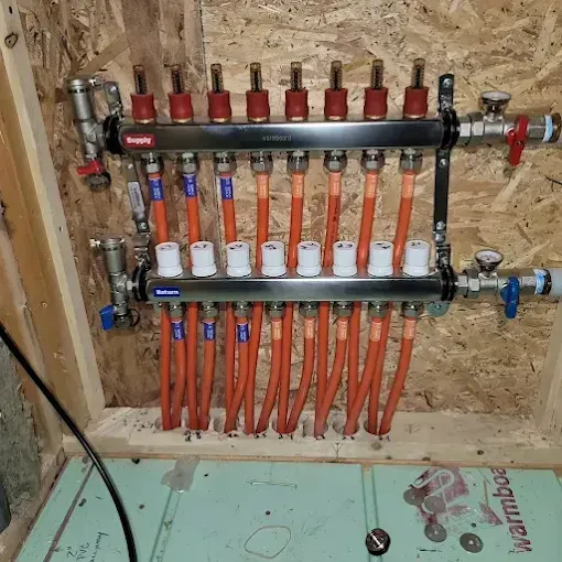 A hydronic heating manifold with eight orange pipes connected to silver bars against an oriented strand board wall.