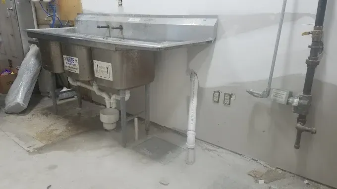 A three-compartment stainless steel utility sink stands against a wall next to exposed plumbing pipes and electrical outlets.