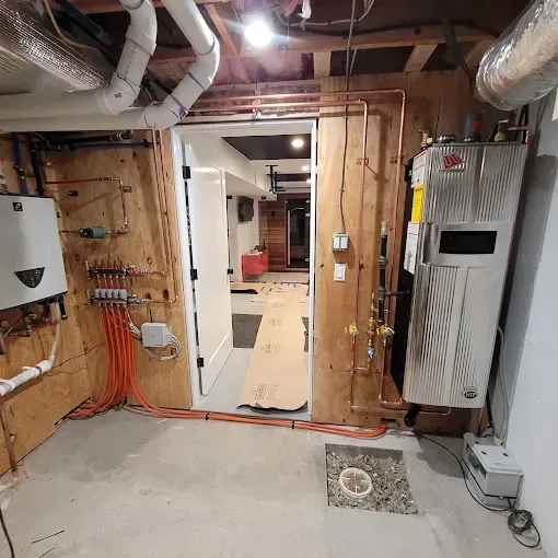 A basement utility room with a white boiler, copper pipes, orange floor heating tubes, and a doorway to a hallway.