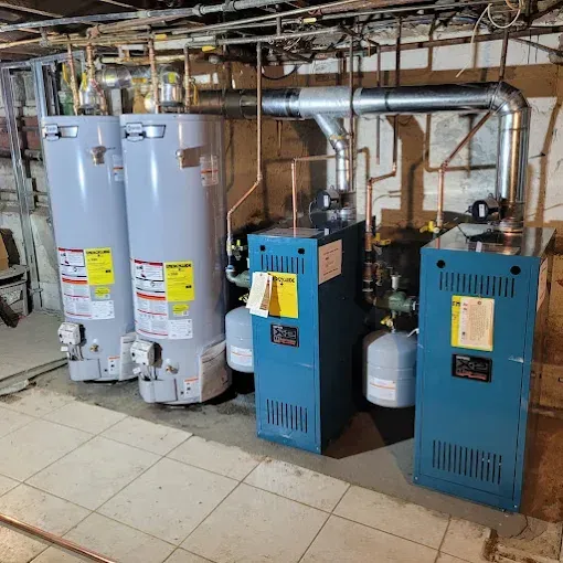 Two grey water heaters and two blue boilers connected to pipes and silver venting ducts in a basement.