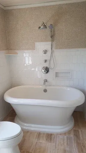 A white freestanding bathtub with a chrome shower fixture against a wall of white subway tile and textured wallpaper.