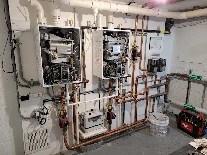 Two wall-mounted HVAC units in a utility room with complex copper piping, a condensate pump, and tools on the floor.