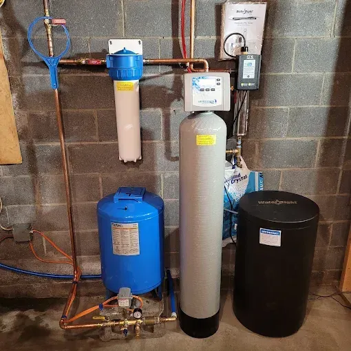 A home water filtration and treatment system with blue and gray tanks, mounted against a concrete block basement wall.