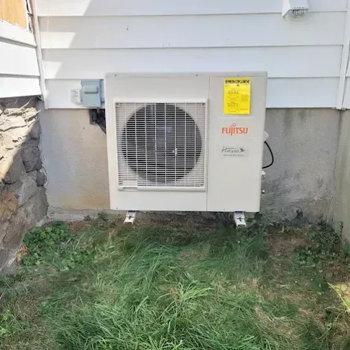 A beige Fujitsu air conditioning unit mounted on a stone wall next to a white house with grass in the foreground.