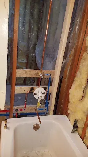 A bathroom under construction showing a bathtub, exposed wooden studs, and new copper and PEX plumbing for a shower faucet.