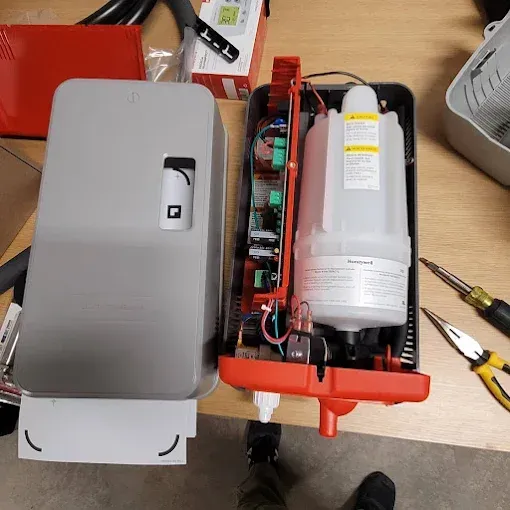 An open wall-mounted soap dispenser casing with its internal reservoir and circuit board visible, alongside tools on a table.