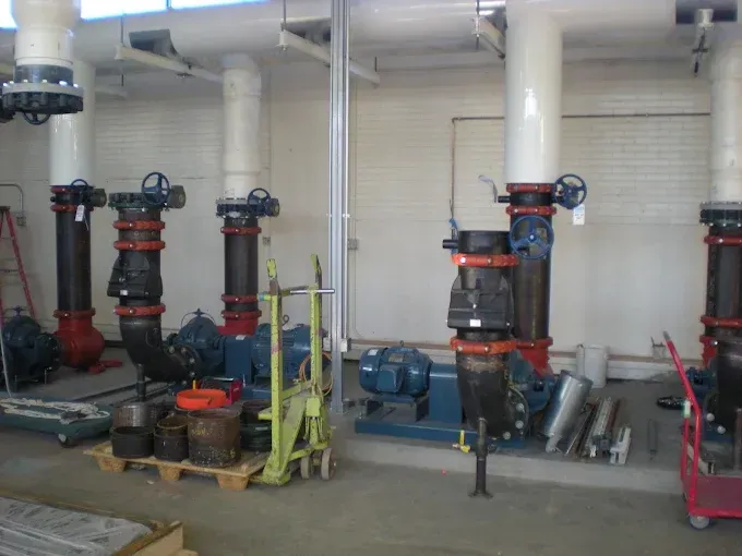 A row of industrial water pumps with black pipes and blue valves in a brightly lit, concrete-floored utility room.