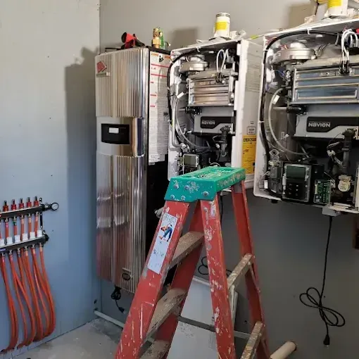 An orange stepladder stands before two wall-mounted Navien tankless water heaters and a radiant heating manifold.