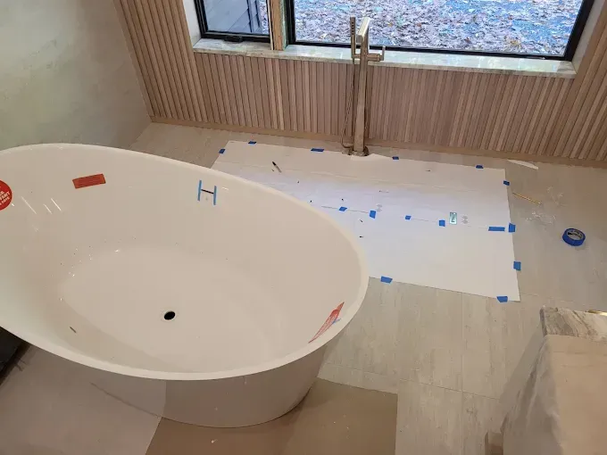 A white freestanding bathtub sits on a tiled floor near a floor-mounted faucet in a room with wood-slat walls.