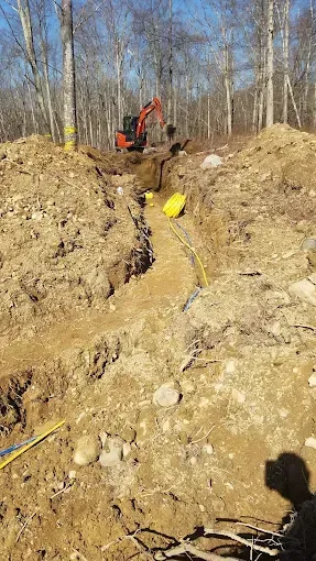 An orange excavator digging a deep dirt trench in a wooded area, with a yellow cable visible along the bottom.