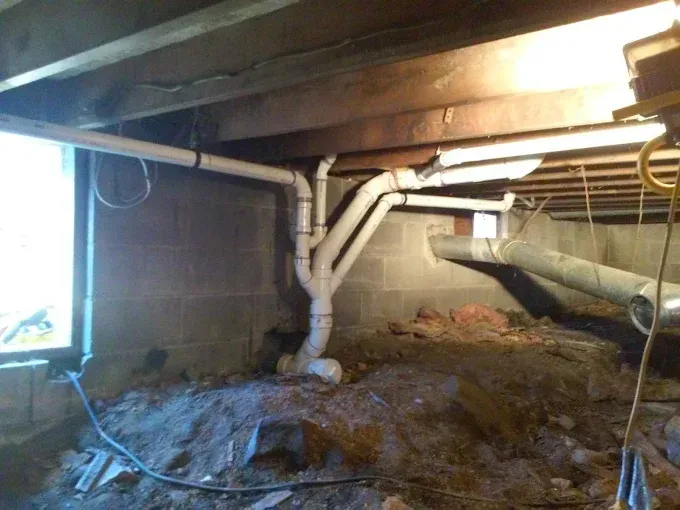 A crawlspace with exposed floor joists, dirt ground, and a complex white PVC plumbing drainage system installed against wall.