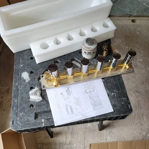 A multi-handle shower valve manifold with brass body and chrome handles sits on a work table next to assembly instructions.